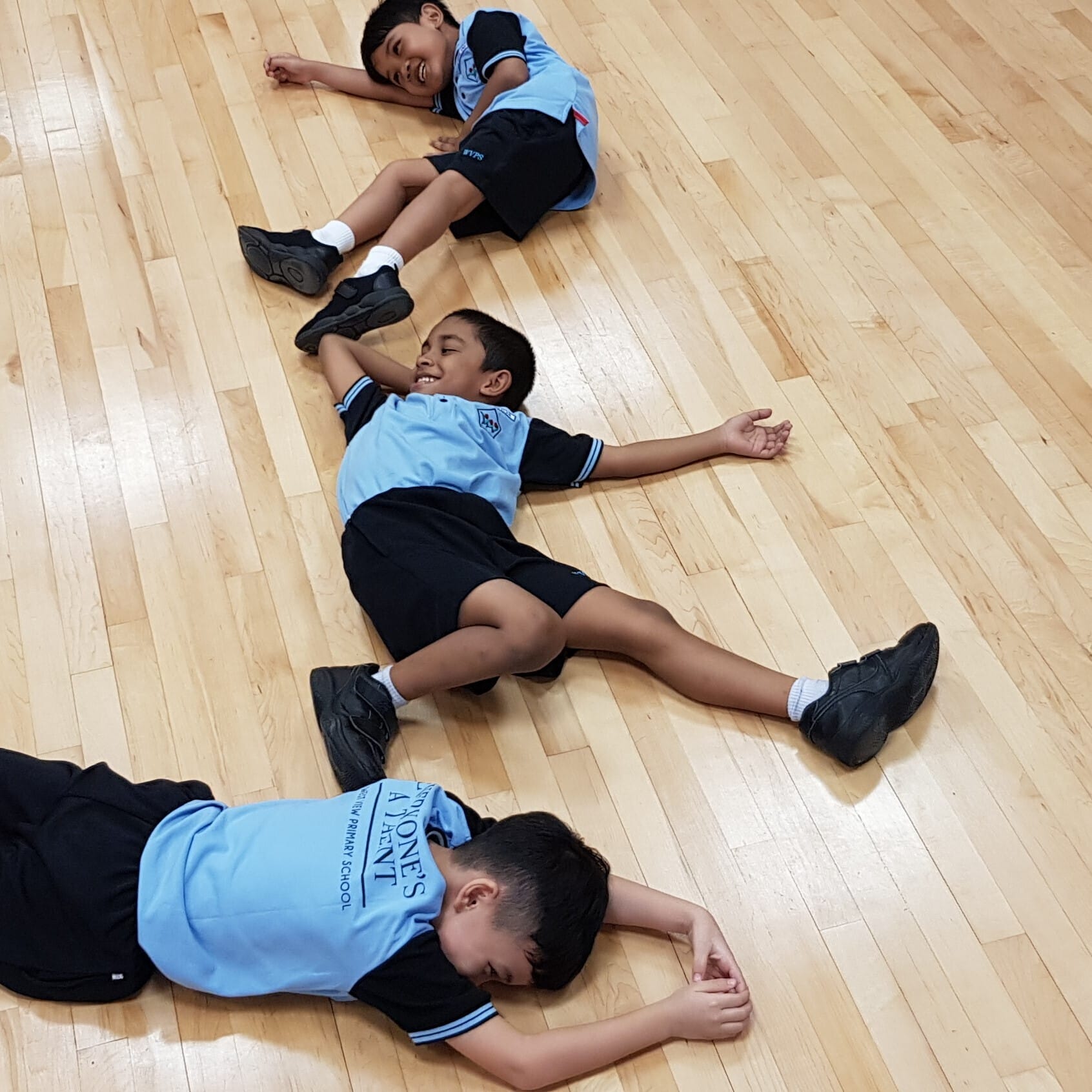 Three boys in blue school uniforms lie and sit on a wooden gym floor, smiling and relaxed.